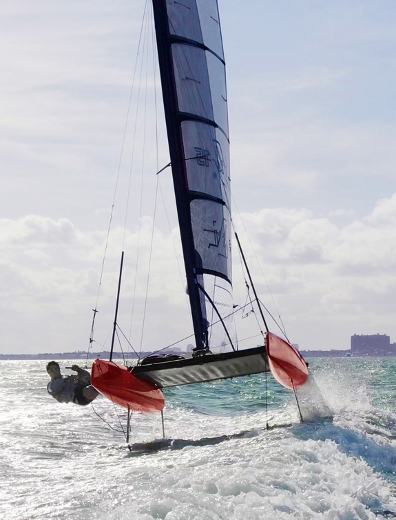 A sailboat with red hulls and a tall sail glides swiftly across choppy water, tilting to one side as two sailors lean out for balance under a bright sky with scattered clouds.
