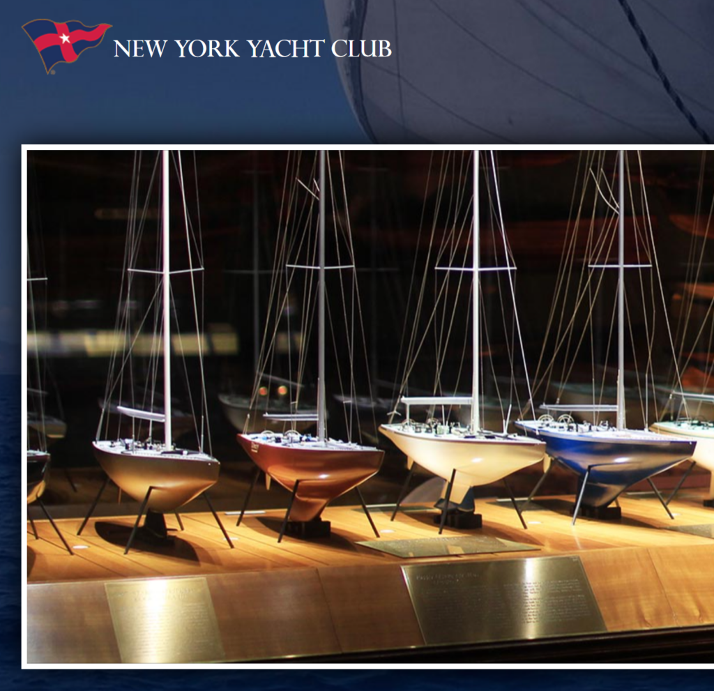 A display of detailed sailboat models with masts and rigging is shown under warm lighting at the New York Yacht Club, with a plaque visible in front of the models. The club’s logo is in the upper left corner.