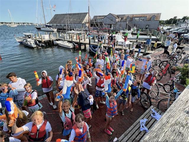 A large group of children and adults, many in life jackets, stand on a dock by sailboats and water, holding up colorful water guns and smiling on a sunny day. Some bikes are parked nearby.