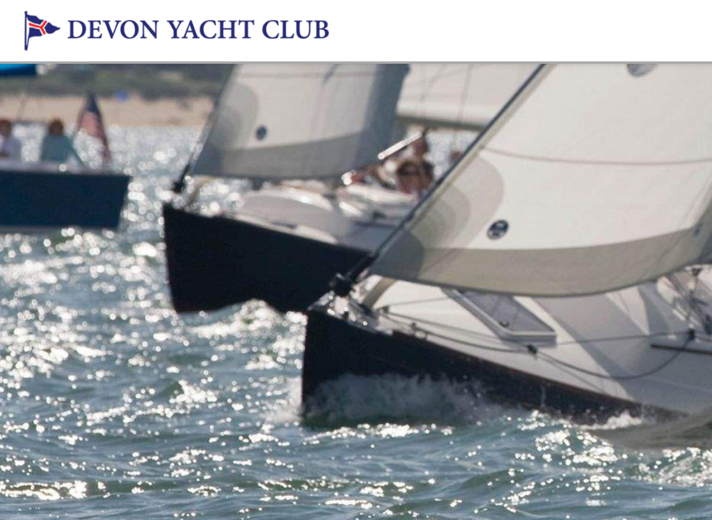 Close-up of two sailboats racing side by side on the water, with their sails full of wind and waves splashing at their bows. The Devon Yacht Club logo and name appear at the top of the image.