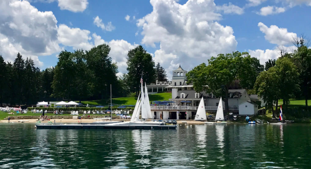 A lakeside scene with sailboats docked near a marina, a sandy beach with people, a large white building, green lawns, trees, and a partly cloudy sky reflected in the calm water.