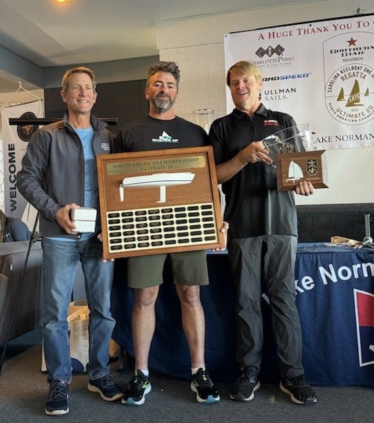 Three men stand indoors, smiling and holding sailing trophies. One holds a large wooden plaque, another has a glass trophy, and the third holds a small box. A blue tablecloth with "Lake Norman" is visible behind them.