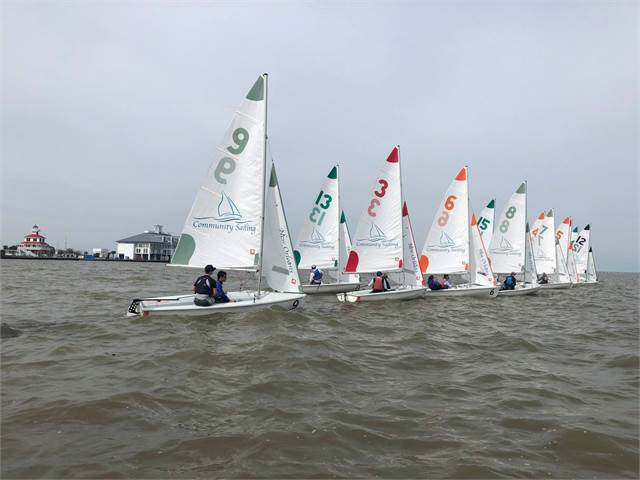 A row of small sailboats with numbered sails, each holding sailors, lines up on choppy water under a cloudy sky, with a building and lighthouse visible in the distance.