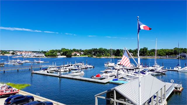 A marina with several docked boats on calm blue water, surrounded by greenery. Two flags, including the American flag, are flying near a small dock office under a clear, bright blue sky.