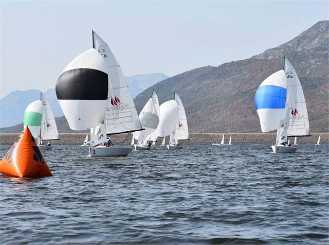 Several sailboats with colorful spinnakers race on a lake near a buoy, with mountains and hills in the background under a clear sky.