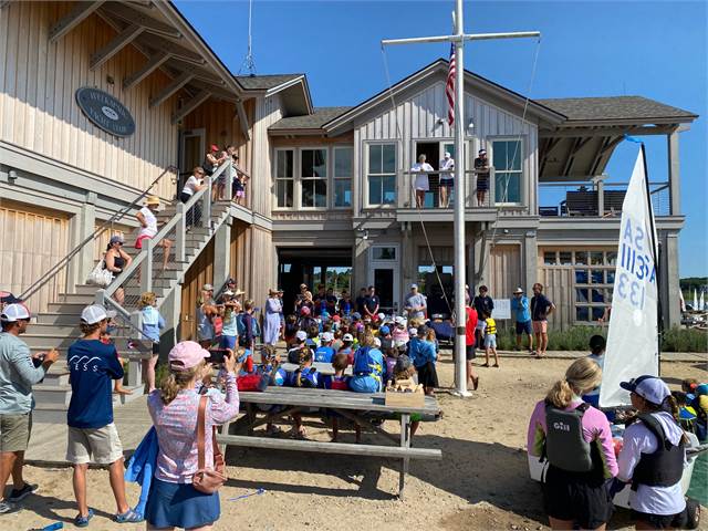 A large group of people, including children and adults, gather outside a wooden building by the water. Some sit on benches, others stand or sit on stairs, while instructors address the group. A sailboat and flagpole are nearby.