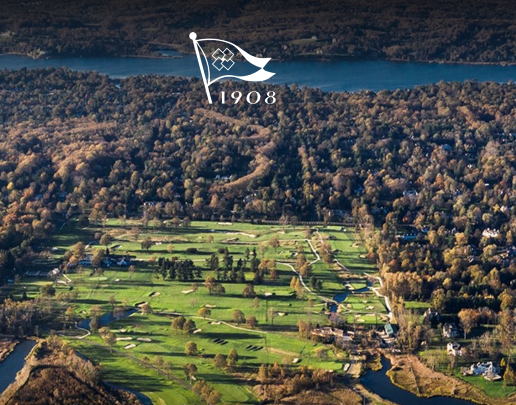 Aerial view of a large golf course surrounded by dense trees and water, with a white flag logo and “1908” superimposed near the top.