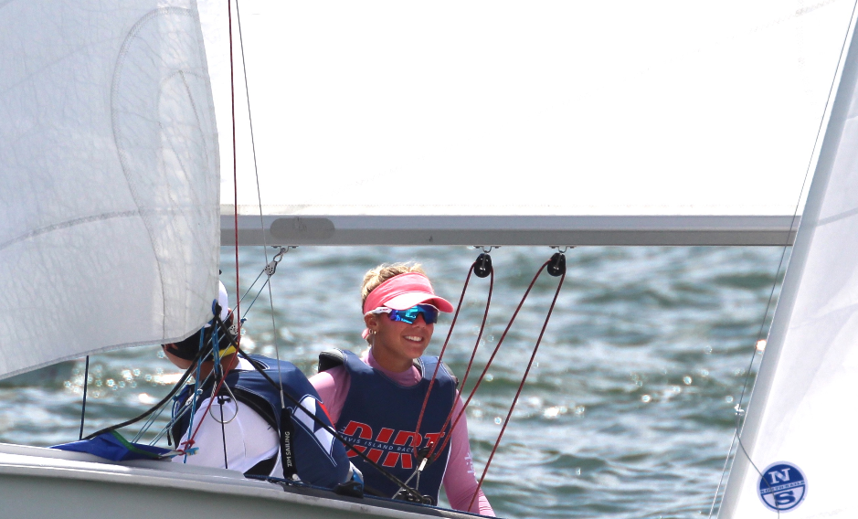 Two people are sailing a small boat on the water. The person in front is smiling, wearing a pink visor, blue sunglasses, and a life vest. White sails and water are visible in the background.