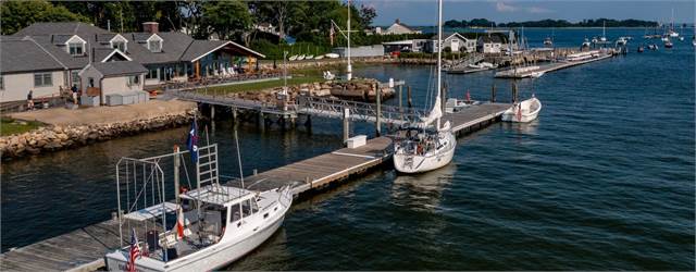 A marina with several boats docked along wooden piers, next to a shoreline with buildings, trees, and outdoor seating areas, under a clear blue sky.