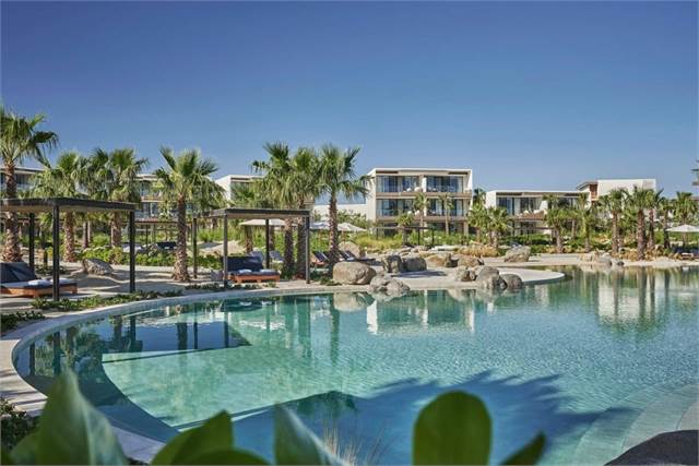 A luxurious resort features a large, clear swimming pool surrounded by palm trees, shaded lounge areas, and modern multi-story buildings under a bright blue sky.