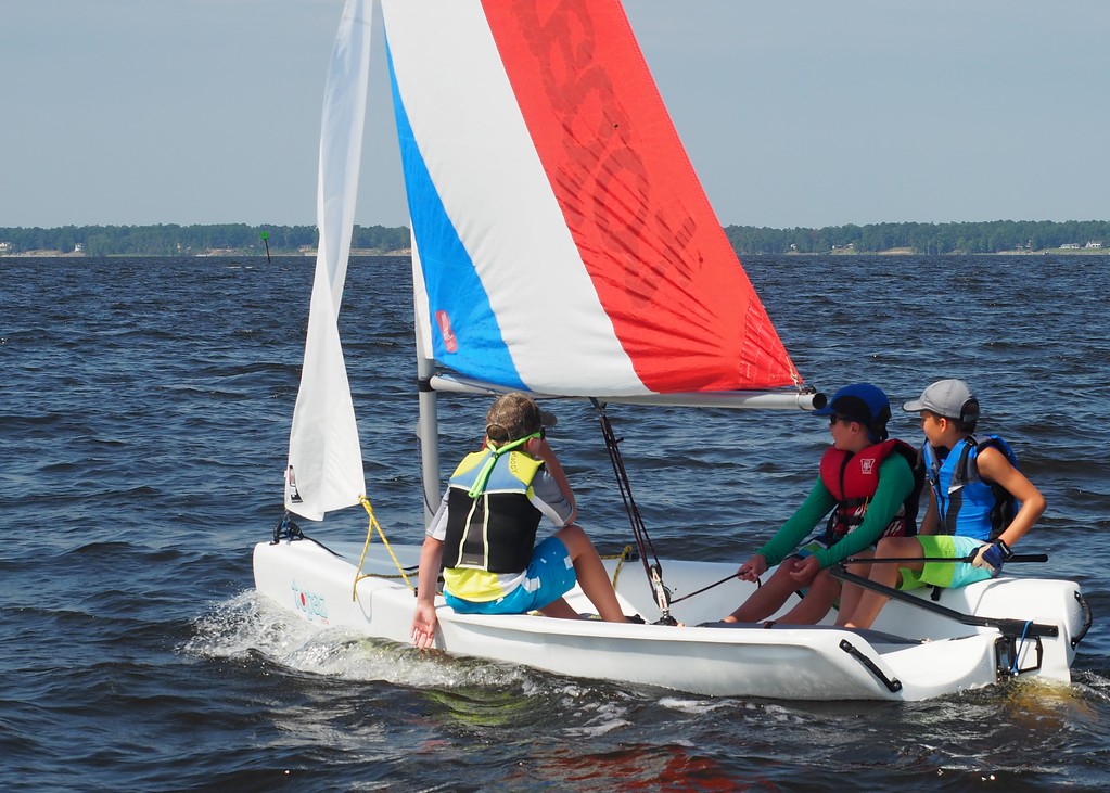 Three children wearing life jackets sail a small boat with a colorful red, white, and blue sail on a lake, with a forested shoreline visible in the background under a clear sky.