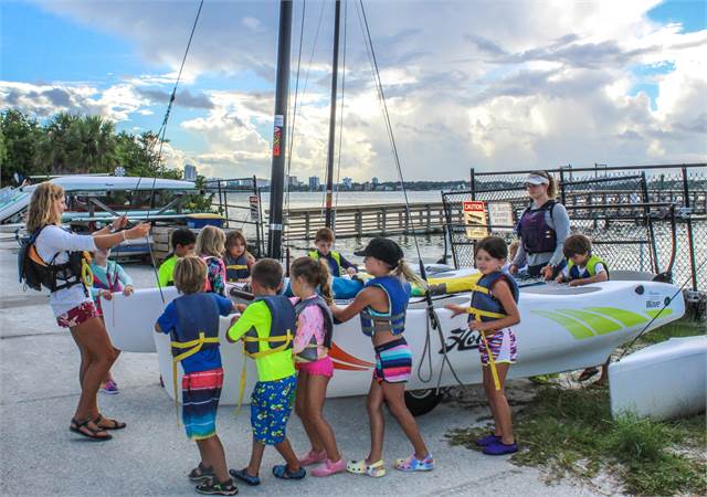 A group of children in colorful life jackets gather around a sailboat as an instructor gives directions near a waterfront on a sunny day. Another adult stands beside the boat, and the water and cityscape are visible in the background.