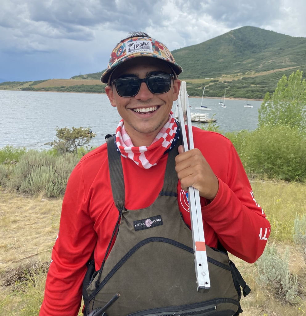A smiling person wearing sunglasses, a colorful cap, and a red shirt stands outdoors by a lake, holding white rods, with hills, boats, and cloudy sky in the background.
