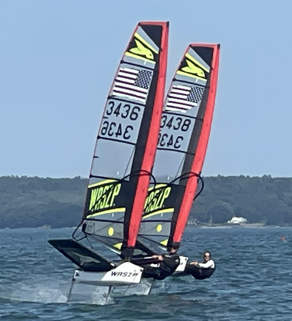 Two people sail side by side on WASZP dinghies with red and yellow sails, each featuring American flags and the numbers 3436 and 3438, on a calm lake with a forested shoreline in the background.