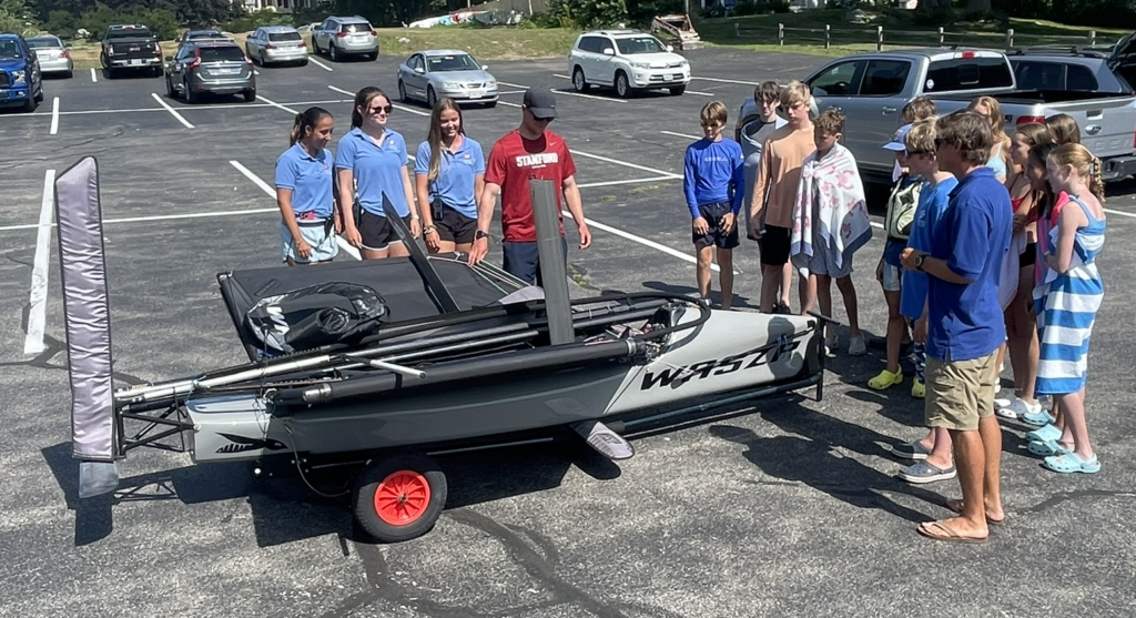 A group of children and teens stand in a parking lot, gathered around a small sailboat on a cart, while an instructor in a red shirt speaks to them. Several cars are parked in the background.