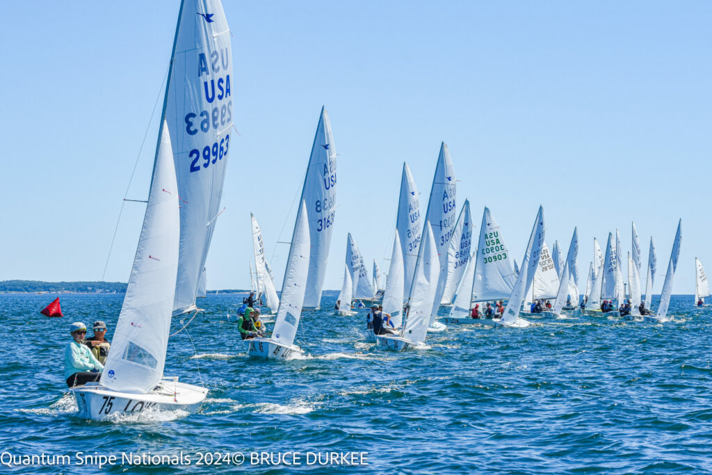 A fleet of small sailboats with white sails races across blue water under a clear sky during the Quantum Snipe Nationals 2024. Sailors are visible on each boat, with land faintly seen in the background.
