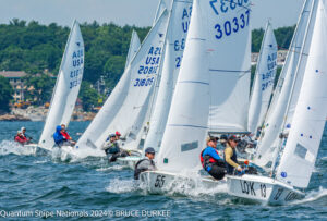 Sailboats with crews compete in a close race on choppy water, sails marked with numbers and “USA.” Trees and buildings line the distant shore under a blue sky.