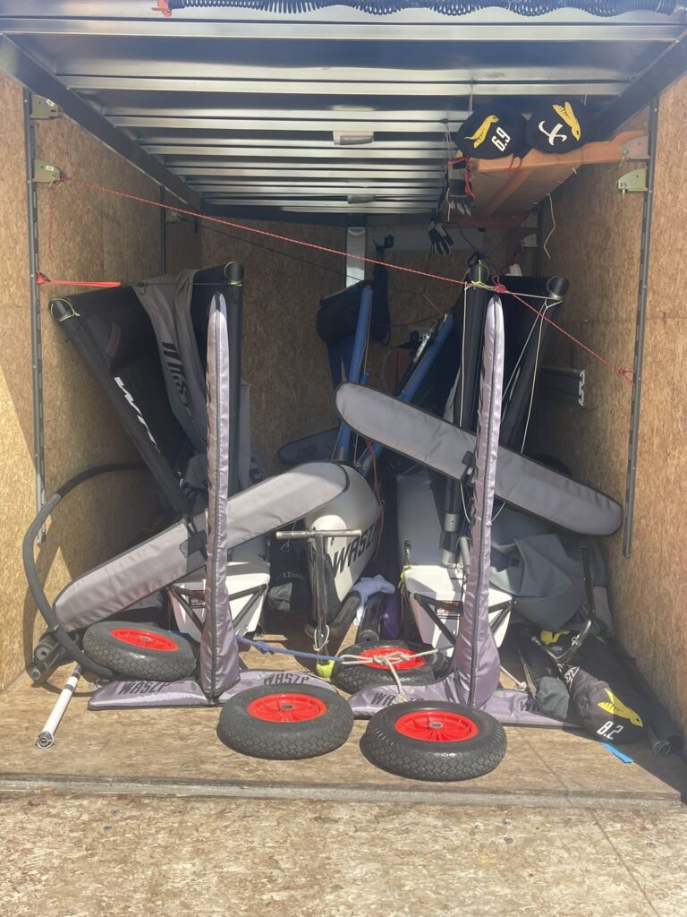 Three small sailboats with gray sails and red-wheeled trolleys are tightly packed in a wooden-walled trailer, with sailing gear and equipment arranged around them. Sunlight shines in from the open trailer door.