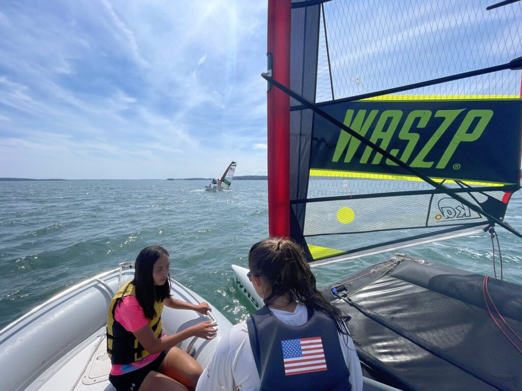 Two people wearing life vests sit on a boat in sunny weather, with one person facing the water. A WASZP sailboat and another sailing vessel are visible on the blue water under a partly cloudy sky.