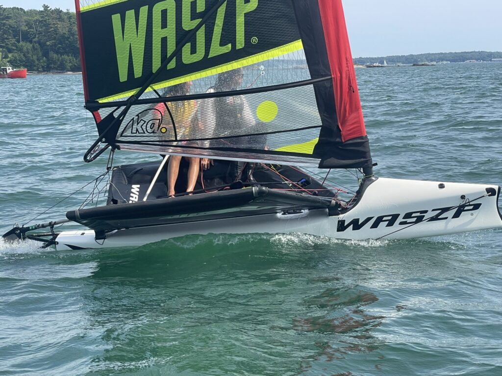 A close-up of a small white WASZP sailboat gliding on green-blue water, with two people on board. The sail is black with yellow and green accents. Trees and another boat are visible in the background.