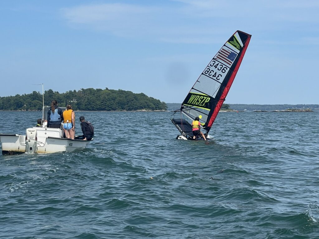 A group of people on a motorboat watch a person windsurfing nearby on a sunny day with an island and trees in the background. The water is slightly choppy, and the sky is mostly clear.