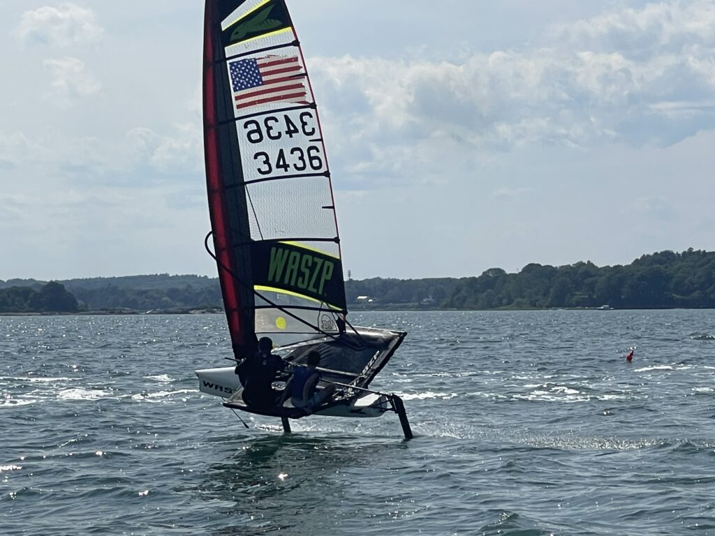 A person sails a small, single-handed hydrofoil sailboat labeled "WASZP" with the number 3436 on its sail, gliding above the water on a sunny day with trees and hills in the background.