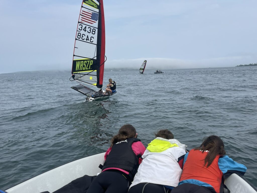 Three children in life jackets lean over the edge of a boat, watching a small sailboat with a colorful sail and an American flag out on a lake. Another sailboat is visible in the distance under a cloudy sky.