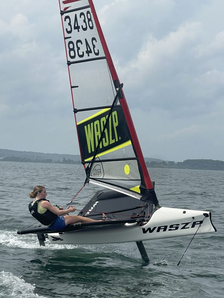 A person sails a white hydrofoil dinghy with "WASZP" written on the sail and hull across a lake, leaning back and holding the ropes, with water splashing beneath the boat and a cloudy sky overhead.