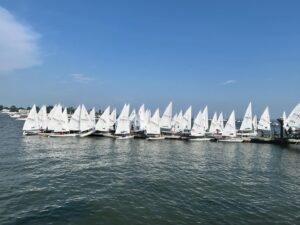 A row of small sailboats with white sails are lined up along a dock on a sunny day, with calm water and a clear blue sky in the background.