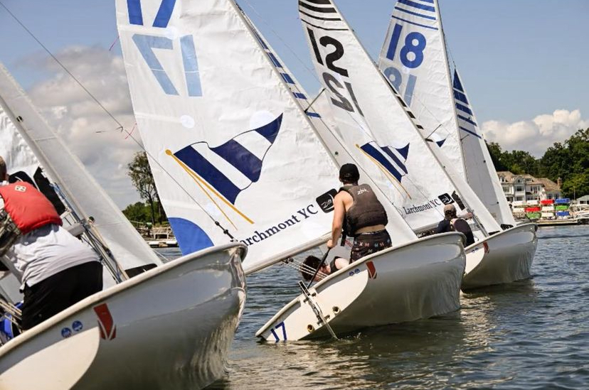Sailboats with numbered white sails race closely on a sunny day, with sailors leaning out over the water. Trees and waterfront houses are visible in the background.