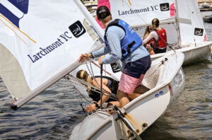 Three people prepare a small sailboat marked "Larchmont YC" at a dock. One person in a blue life vest stands, while two others adjust sails and lines. Other similar boats are visible nearby on the water.