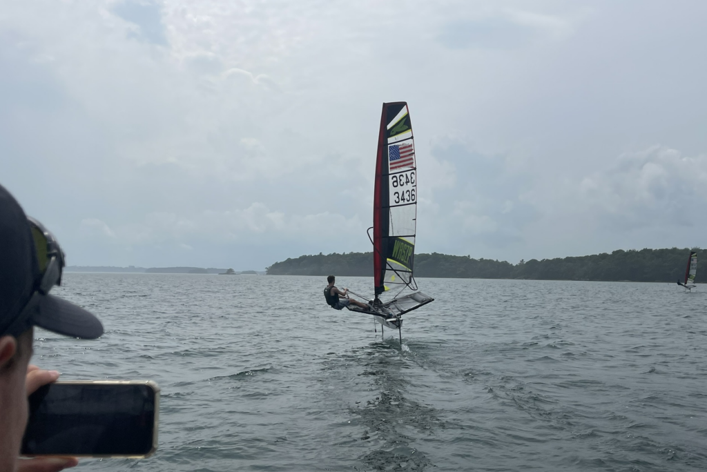 A person sails a windfoiling board over the water on a cloudy day. Another person in the foreground captures the scene on their phone. A small tree-covered island is visible in the distance.