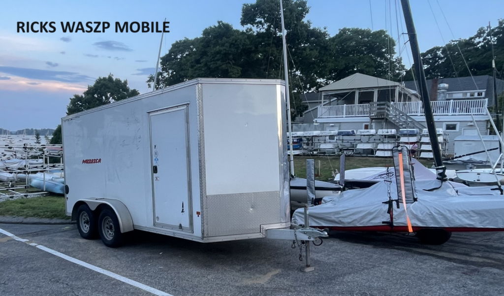 A white enclosed trailer labeled "RICK'S WASZP MOBILE" is parked on a paved lot near docks with several small sailboats and trees in the background, during early evening.