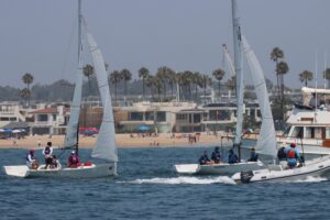 Two small sailboats with crews racing near a beach, with a motorboat nearby. People are visible on the sandy beach and palm trees and buildings line the shore in the background.