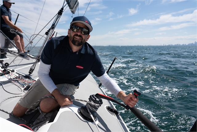 A man wearing sunglasses and a cap is smiling while sailing a boat on a sunny day. He is gripping a winch handle, with the open sea and city skyline visible in the background.