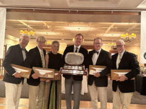 Six people dressed in formal attire stand indoors, smiling and holding wooden sailing awards. The person in the center holds a large silver trophy on a wooden base. Chandeliers and tables are visible in the background.