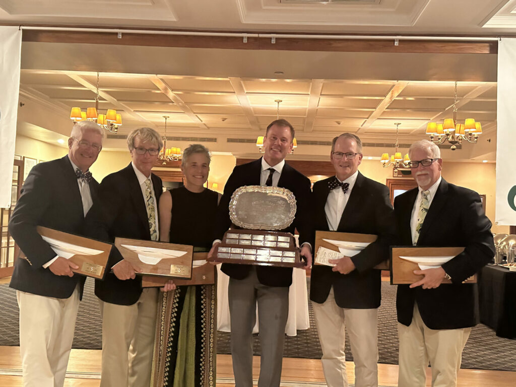 Six people dressed in formal attire stand indoors, smiling and holding wooden sailing awards. The person in the center holds a large silver trophy on a wooden base. Chandeliers and tables are visible in the background.