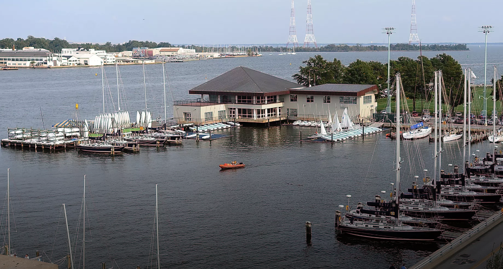 A marina with sailboats docked around a large, modern building on the water, some boats moored and others in the harbor. A small orange boat is on the water, and there are radio towers in the background.