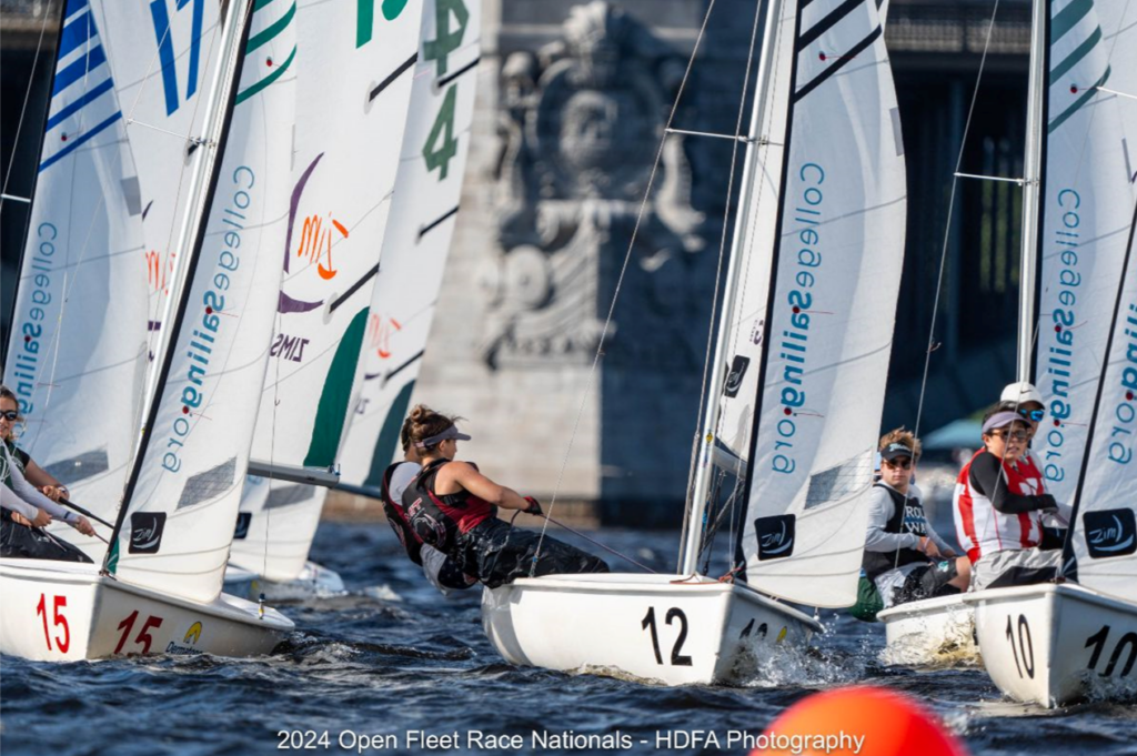 Sailboats with college sailors compete closely in a regatta, racing in choppy water near a stone bridge. Sail numbers and the word 'Collegesailing.org' are visible on the sails.