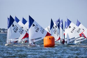 A group of small sailboats with white, blue, and red sails race around an orange buoy on a calm body of water, with sailors visible in each boat and a shoreline in the background.