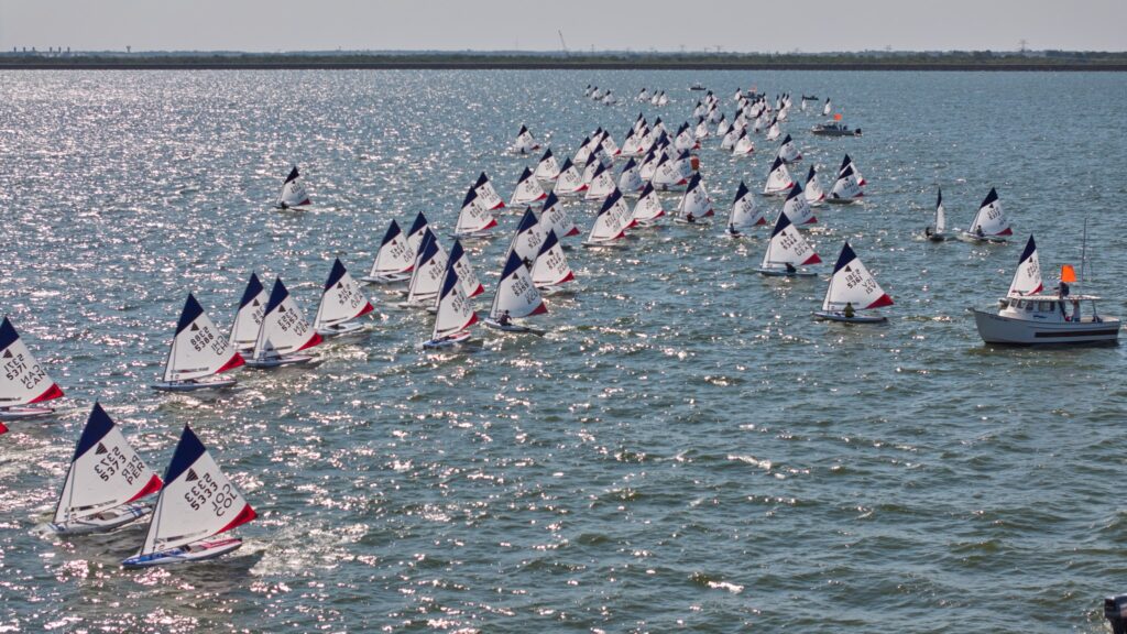 A large group of small sailboats with white sails race across a sunlit body of water, closely aligned in rows, with a motorboat on the right displaying an orange flag.