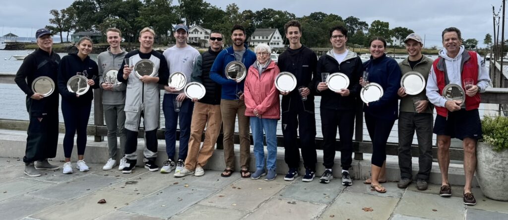 A group of twelve people stands outdoors by a waterfront, smiling and holding silver plates and glass awards. Trees and houses are visible in the background under a cloudy sky.