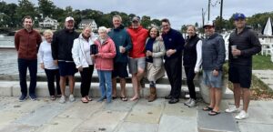 A group of thirteen people stand close together, smiling outdoors on a stone walkway by the water. Trees and houses are visible in the background on a cloudy day. Some people hold drinks; one holds a small plaque.