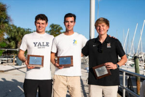 Three young men stand side by side outdoors, each holding a plaque. They are smiling, with sailboats and palm trees in the background. The left person wears a "Yale Sailing" shirt, the middle a white tee, and the right a black polo.