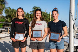 Three young women stand side by side outdoors, smiling and holding plaques. One wears a Cornell Sailing shirt, another a Harvard shirt, and the third a dark shirt, with palm trees and boats visible in the background.