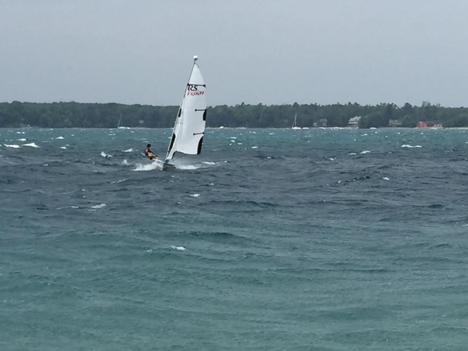 A small sailboat with one person aboard sails on a choppy, windy lake. Whitecaps are visible on the dark blue water, and a forested shoreline appears in the background under a cloudy sky.