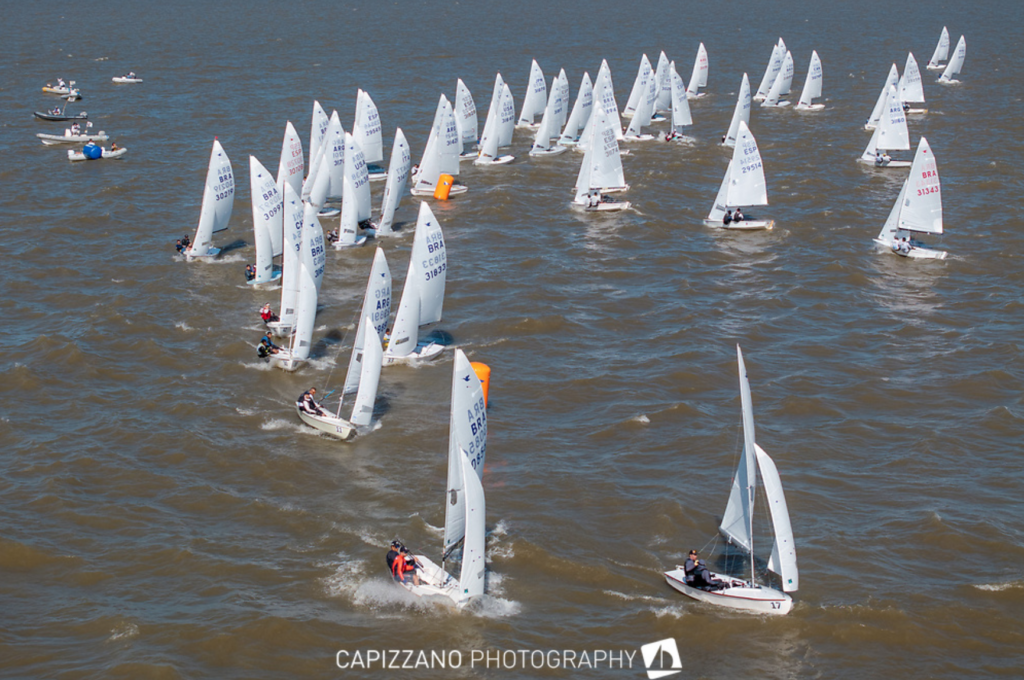 A large group of small sailboats with white sails race on choppy blue water. Some boats turn around an orange buoy while others follow behind. Several people are visible in each boat under a clear sky.
