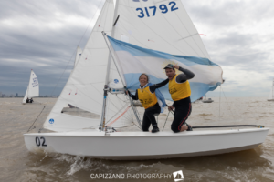 Two sailors in yellow vests smile and wave on a small sailboat with the number 02, holding an Argentine flag. Other sailboats are visible on the water under a cloudy sky.