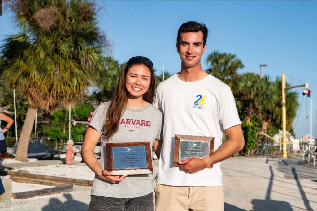 Two young adults stand outdoors holding plaques and smiling. The woman wears a gray “Harvard Sailing” shirt, while the man wears a white shirt with a blue and yellow logo. Palm trees and boats are visible in the background.
