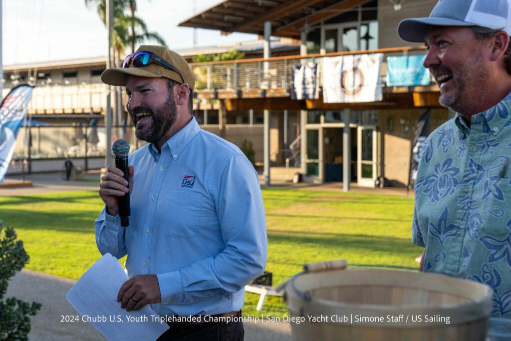 A man in a light blue shirt and brown cap speaks into a microphone while holding papers, standing outdoors near another smiling man, with a modern building and palm trees in the background.
